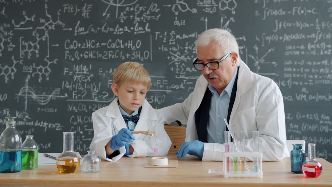 Chemistry professor and little child doing chemical test in school laboratory holding glass tube talking sitting at desk with chalkboard wall in background.