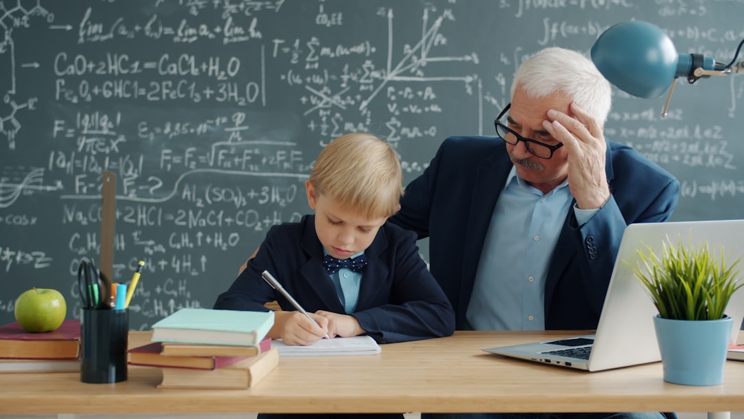Little boy studying with tutor writing at desk in classroom focused on education, black chalkboard with formulas is visible in background. People and science concept.